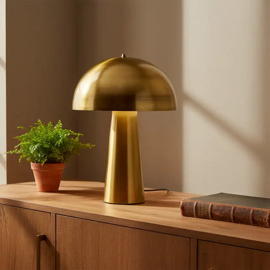 Gold table lamp on a wooden surface with a plant and book in a warm setting