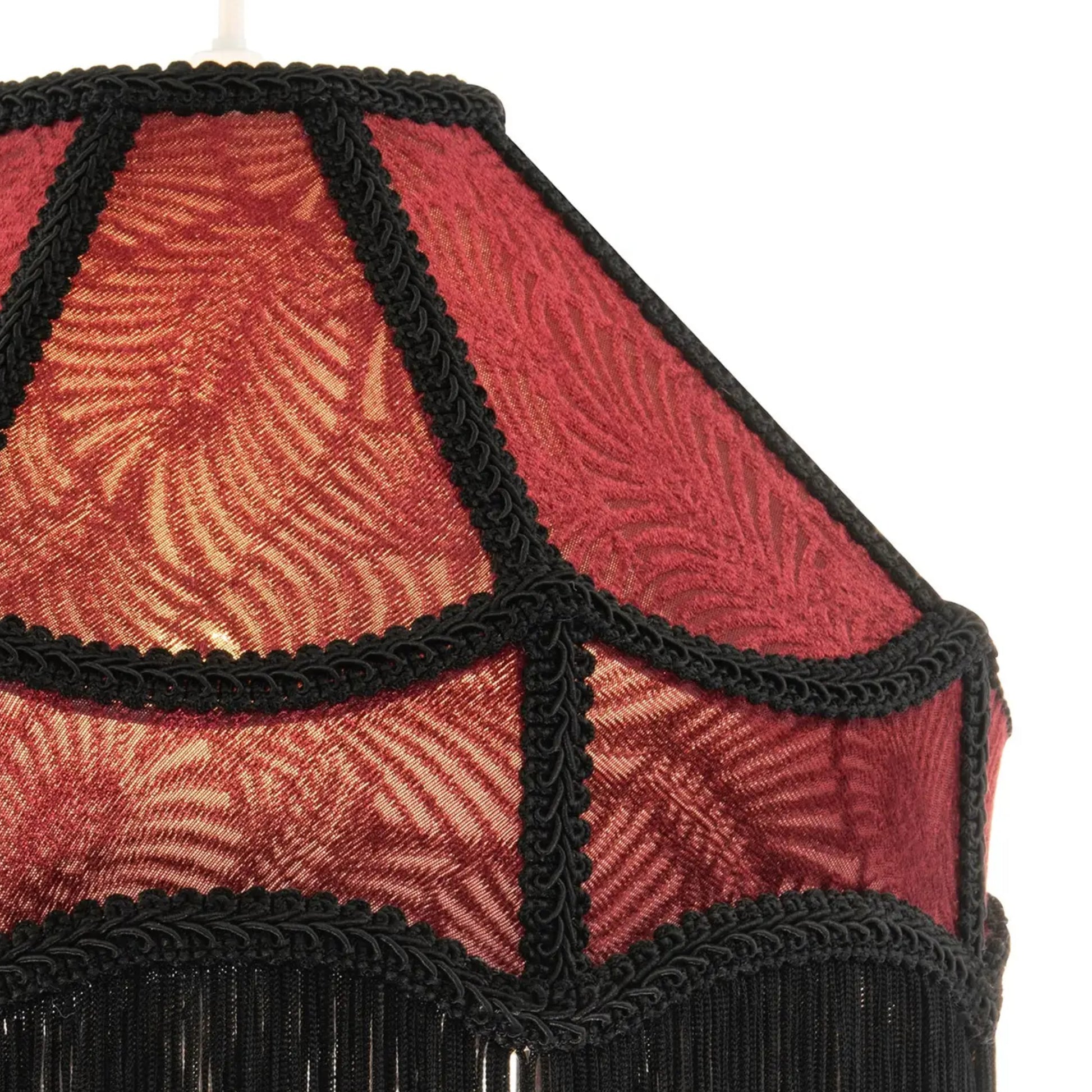 Close-up of a decorative lampshade with a red and gold leaf patterned fabric and black braided trim with fringe at the bottom, set against a plain white background.