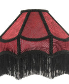 A vintage-style fabric lampshade with a red patterned body, black braided trim, and a long black fringe along the bottom edge, shown against a plain white background.