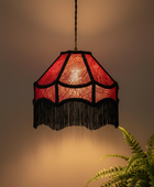 A hanging pendant lamp with a red patterned shade and black fringe, suspended against a warm beige wall with a green plant visible in the lower right corner.