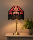 A vintage-style table lamp with a red and black patterned fringed shade and a gold brass stem, sitting on a light-colored wooden table next to a potted fern.
