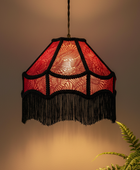A vintage-style pendant lamp with a red patterned fabric shade and black fringe, hanging against a warm beige wall with a green fern in the foreground.