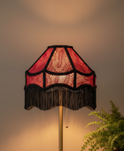 A vintage-style floor lamp with a red and black patterned fringed shade, illuminated to cast a warm glow against a neutral background with a green fern in the foreground.