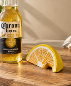 A bottle of Corona Extra beer stands on a wooden cutting board next to a sliced orange wedge, with a blurred kitchen background including a plant, bowl, and cloth.