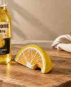 A ceramic orange slice candle holder sits on a wooden table next to a Corona Extra bottle, with a neutral background and a folded cloth.