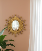 Decorative gold sunburst mirror on a beige wall with a plant and window in the foreground.