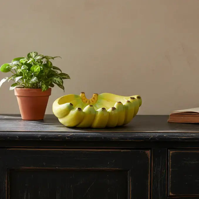 Decorative fruit bowl shaped like a banana on a wooden surface with a plant and book in the background.