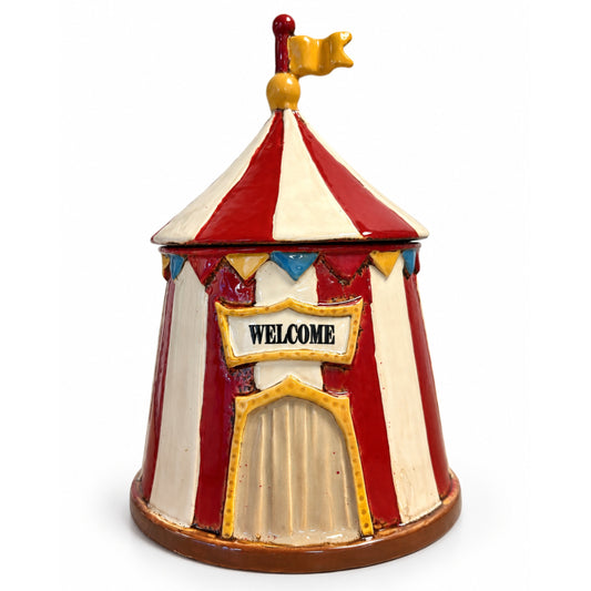 A small circus tent shaped container or decorative piece with red and cream vertical stripes, a gold-trimmed front door, a 'WELCOME' sign, and a gold flag on top, photographed on a plain white background.
