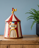 A ceramic circus tent cookie or decorative container with red and white stripes, a yellow 'WELCOME' sign, and a small flag on top, displayed on a wooden cabinet next to a potted fern against a blue wall.