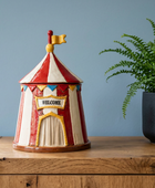A ceramic circus tent-shaped container with a red and white striped pattern, a yellow 'WELCOME' sign, and a small yellow flag on top, displayed on a wooden surface next to a potted fern against a blue wall.
