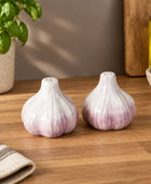 Two garlic-shaped salt and pepper shakers with a glossy white and purple glaze, set on a white tablecloth with a glass vase, tomatoes, and a wooden cutting board in the background.