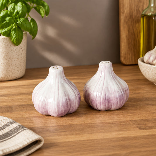 Two garlic-shaped salt and pepper shakers with a glossy white and purple glaze, set on a white tablecloth with a glass vase, tomatoes, and a wooden cutting board in the background.