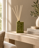 An olive green reed diffuser bottle with embossed text and several natural reed sticks, styled on a light wood table in a warm, sunlit room next to a sofa and books.