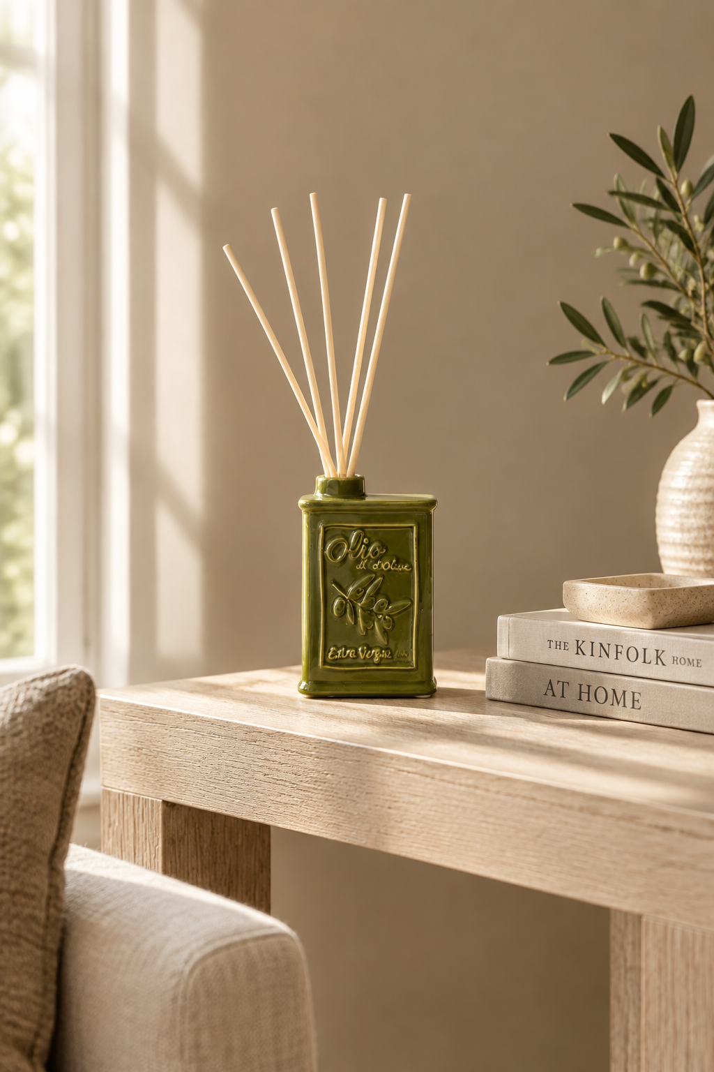 An olive green reed diffuser bottle with embossed text and several natural reed sticks, styled on a light wood table in a warm, sunlit room next to a sofa and books.