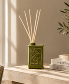 An olive green reed diffuser bottle with embossed olive branch details and natural reed sticks, styled on a light wood table next to stacked books and a vase in a softly lit room.