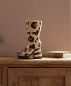 Decorative boot with leopard print on a wooden surface next to a plant and book.