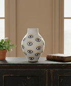 Decorative vase with eye pattern on a wooden surface with a plant and book in the background.