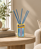 A reed diffuser styled on a round wooden table in a cozy room setting. The diffuser has a yellow and light blue fish-shaped container with the word 'SARDINE' printed on it, and four blue reeds extending upwards.