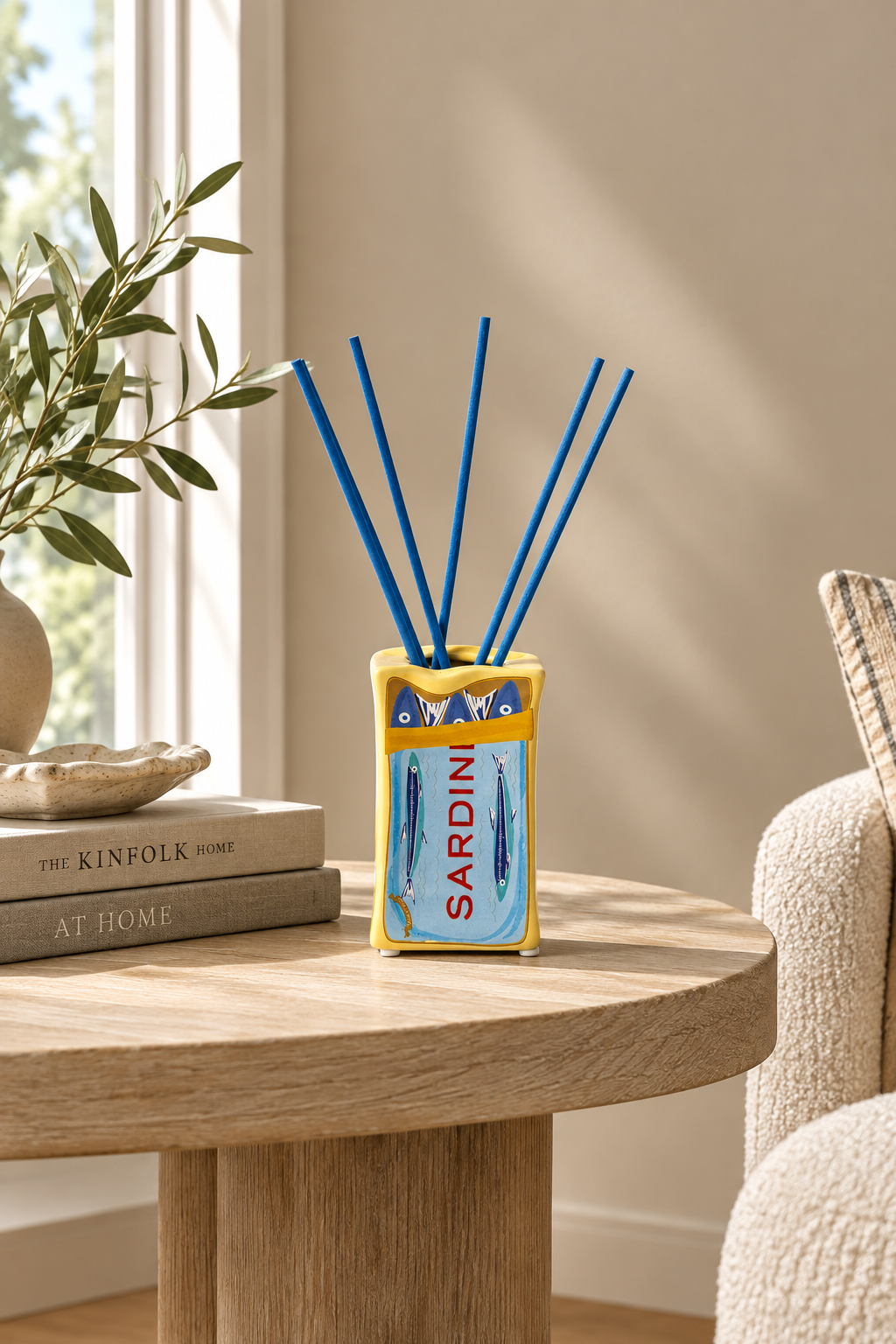A reed diffuser styled on a round wooden table in a cozy room setting. The diffuser has a yellow and light blue fish-shaped container with the word 'SARDINE' printed on it, and four blue reeds extending upwards.