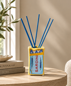 A decorative reed diffuser vase with blue reeds, placed on a round wooden table in a styled interior. The vase has a yellow border and a blue front panel with fish illustrations and the word 'SARDINI' in red.