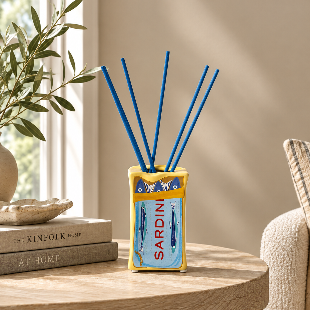 A decorative reed diffuser vase with blue reeds, placed on a round wooden table in a styled interior. The vase has a yellow border and a blue front panel with fish illustrations and the word 'SARDINI' in red.