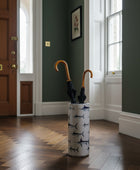 A cylindrical umbrella stand with a beige background and a repeating print of sailfish, holding two closed black umbrellas with curved orange handles, set against a plain white background.