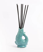 A turquoise ceramic diffuser shaped like a stylized fish or creature, holding six black reeds, photographed on a clean white background.