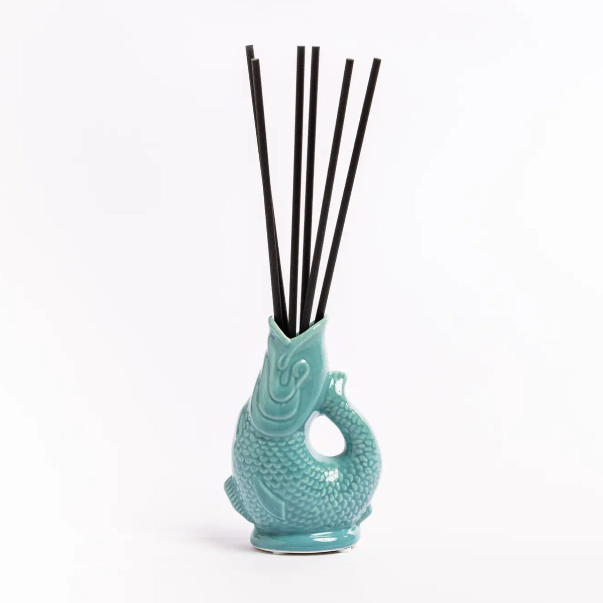 A turquoise ceramic diffuser shaped like a stylized fish or creature, holding six black reeds, photographed on a clean white background.