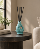 A turquoise ceramic fish-shaped reed diffuser with black reeds on a round dark wood table in a softly lit room, with a chair and books partially visible.