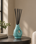 A turquoise ceramic fish-shaped reed diffuser with black reeds, displayed on a dark wooden table in a cozy, sunlit room next to a potted plant and a beige armchair.