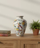 Decorative vase with floral and parrot design on a wooden surface with books and a plant.