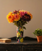 Colorful vase with flowers on a wooden surface next to a book and small potted plant.
