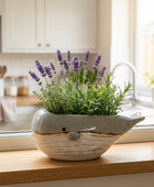 A whale-shaped ceramic planter with a light blue-gray top and a textured off-white lower body, filled with artificial green foliage and small purple flowers, displayed on a round wooden tray.