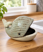 A ceramic whale-shaped planter with a light blue-green glazed exterior and a beige interior, placed on a wooden kitchen countertop next to a potted basil plant and a white sink.