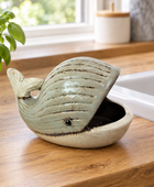 A whale-shaped ceramic planter with a light blue-green glazed exterior and a beige interior, shown on a wooden countertop near a sink and a potted plant.