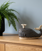 A dark glazed ceramic whale-shaped lidded box with a cream-colored top and tail, displayed on a wooden sideboard next to a potted green fern against a light blue-gray wall.