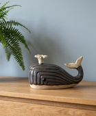 A ceramic whale-shaped lidded container with a dark brown and beige glaze, displayed on a wooden sideboard next to a green fern plant against a blue-gray wall.
