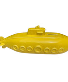 A bright yellow submarine-shaped decorative object, possibly a pig bank or money box, displayed on a light marble surface with potted plants in the background.