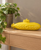 A bright yellow submarine-shaped decorative object resting on a light wood console table, with a potted green plant and a beige wall in the background.