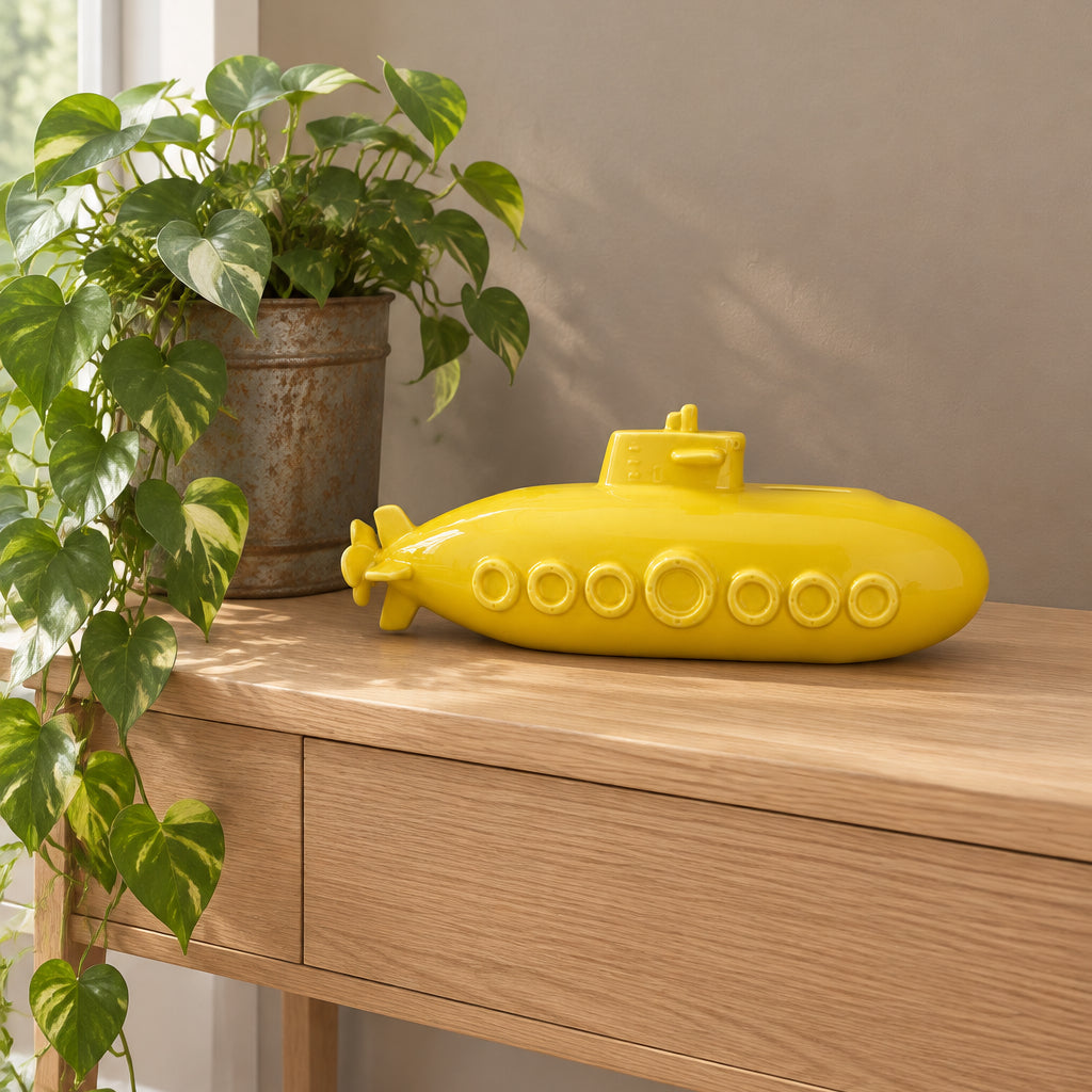 A bright yellow submarine-shaped decorative object resting on a light wood console table, with a potted green plant and a beige wall in the background.