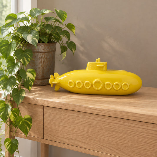 A bright yellow submarine-shaped decorative object resting on a light wood console table, with a potted green plant and a beige wall in the background.