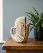 A ceramic fish-shaped planter with a curved handle and spout, featuring textured concentric ridges and a small black eye detail, sitting on a wooden table next to a potted green fern.