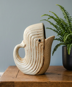 A ceramic whale-shaped pitcher with a cream-colored glaze and brown speckled detailing, styled on a wooden table next to a potted fern against a blue-gray wall.