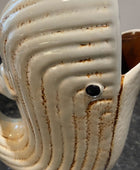 A close-up of a ceramic whale-shaped pitcher with a glossy off-white glaze, brown distressed streaks, and a small black eye detail, set on a dark countertop.