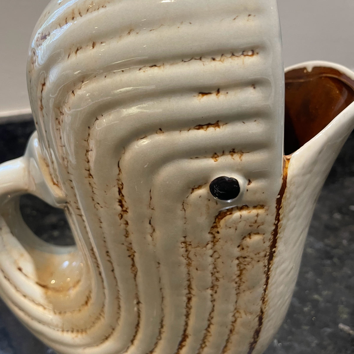 A close-up of a ceramic whale-shaped pitcher with a glossy off-white glaze, brown distressed streaks, and a small black eye detail, set on a dark countertop.