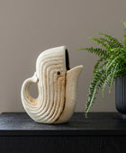 A sculptural ceramic pitcher shaped like a stylized bird, in an off-white glaze with subtle brown weathered accents, displayed on a dark wooden cabinet next to a potted green fern.
