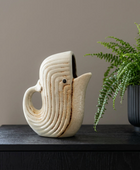 A sculptural ceramic pitcher shaped like a stylized bird or duck, shown on a dark wooden table next to a potted green fern against a neutral beige wall.