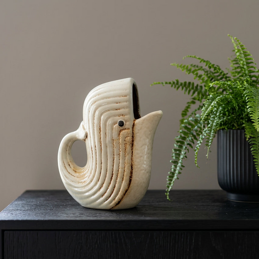 A sculptural ceramic pitcher shaped like a stylized bird or duck, shown on a dark wooden table next to a potted green fern against a neutral beige wall.