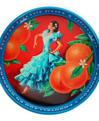 A round metal tin lid featuring a vibrant illustration of a woman in a light blue dress dancing among large oranges and green leaves on a red background, with a blue rim inscribed with Spanish text.