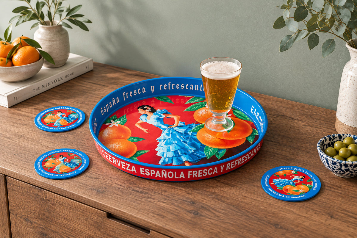 A round decorative serving tray with a colorful Spanish-themed illustration, placed on a rustic wooden table. The tray features a red background with orange fruits, green leaves, a woman in a blue dress, and blue rim accents.